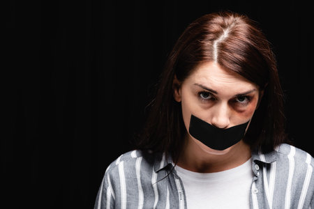Victim of domestic abuse with bruise on face and adhesive tape looking at camera isolated on blackの写真素材