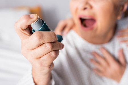 cropped view of senior woman with open mouth holding inhaler while suffering from asthma attack,...