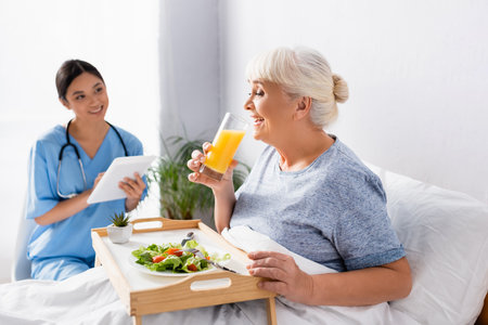 pleased aged woman having breakfast and drinking orange juice near smiling asian nurse on blurred backgroundの写真素材