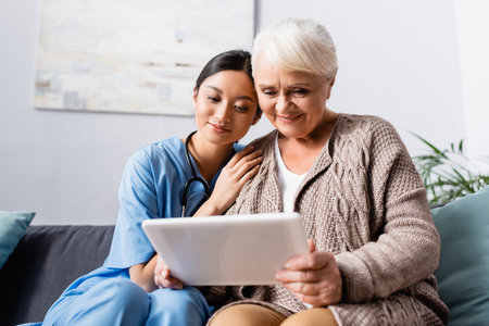 happy asian woman leaning on shoulder of aged smiling woman holding digital tabletの写真素材