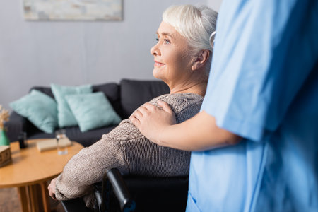 nurse touching shoulder of aged, smiling disabled woman in wheelchair, blurred foreground