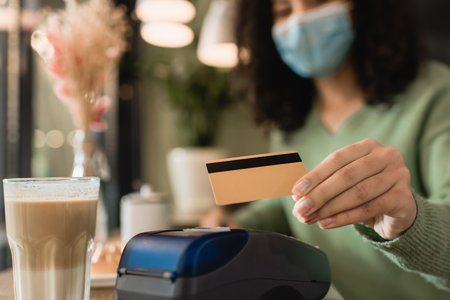glass of latte near credit card in hand of african american woman in medical mask paying on blurred...