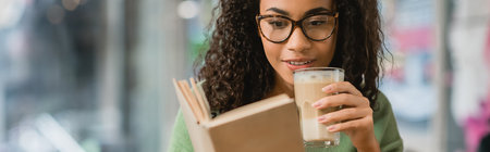 cheerful african american woman reading book and holding glass with latte in cafe, bannerの写真素材