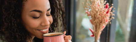pleased african american woman holding pink cup with coffee in cafe, banner