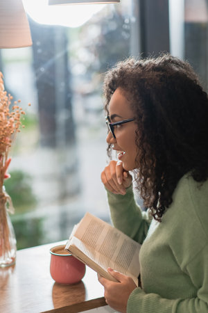 side view of positive african american woman in eyeglasses reading book in cafe