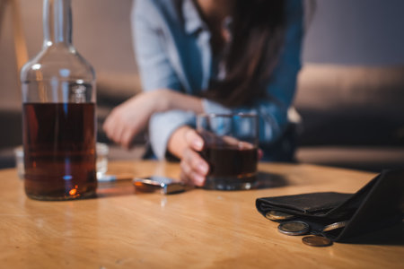 cropped view of alcohol-addicted woman taking glass of whiskey near wallet with coins, blurred backgroundの写真素材
