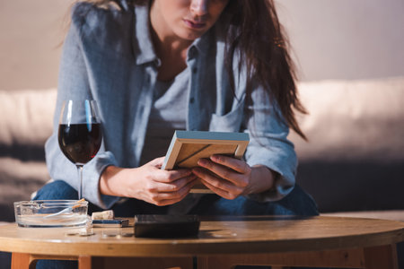 cropped view of depressed woman holding photo frame near glass of red wine, blurred backgroundの写真素材