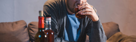 cropped view of alcohol-addicted man drinking whiskey near bottles on blurred foreground, banner