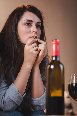frustrated woman holding wedding ring and looking away near bottle and glass of red wine on blurred foregroundの写真素材