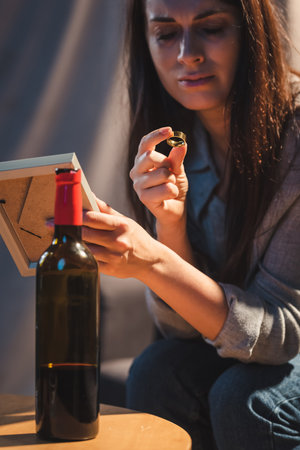 depressed woman holding photo frame and wedding ring near bottle of red wineの写真素材