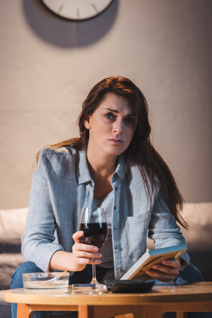 depressed woman looking at camera while sitting with glass of red wine and holding photo frame