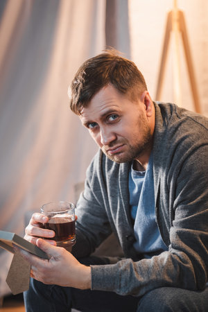 lonely man looking at camera while holding glass of whiskey and photo frameの写真素材