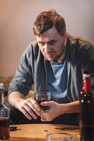 drunk, frustrated man holding glass of whiskey near bottles of alcohol on blurred foregroundの写真素材