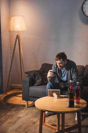depressed man drinking whiskey near table with photo frame and alcohol drinks at home