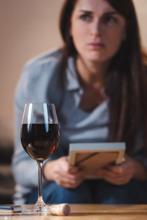 lonely, alcohol-addicted woman holding photo frame while sitting near glass of red wine, blurred backgroundの写真素材