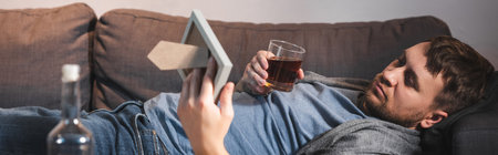 frustrated man lying on sofa with glass of whiskey and photo frame near bottle on blurred foreground, bannerの写真素材