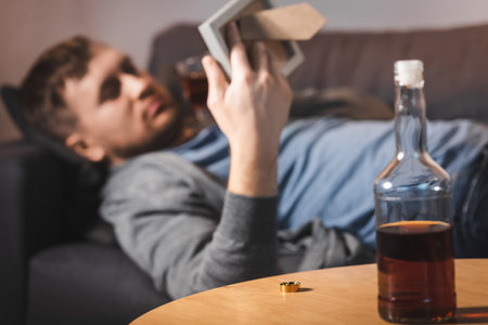 bottle of whiskey and wedding ring on table near depressed man lying on sofa with photo frame on...