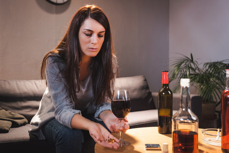 frustrated woman holding wedding ring and glass of red wine near bottles on blurred foregroundの写真素材