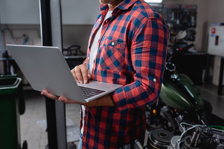 cropped view of technician in plaid shirt with laptop near motorcycle spare parts in garageの写真素材
