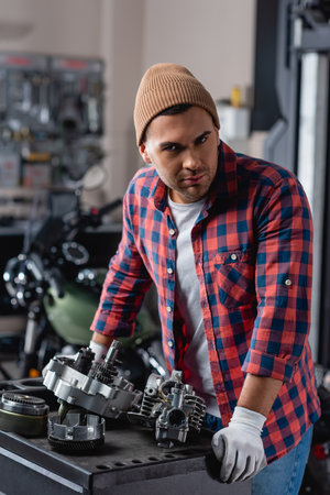 young mechanic in plaid shirt and beanie looking at camera near disassembled motorbike gearboxの写真素材