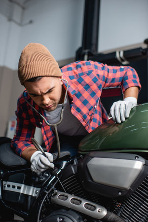 repairman in plaid shirt and hat making diagnostics of motorcycle engine with stethoscopeの写真素材