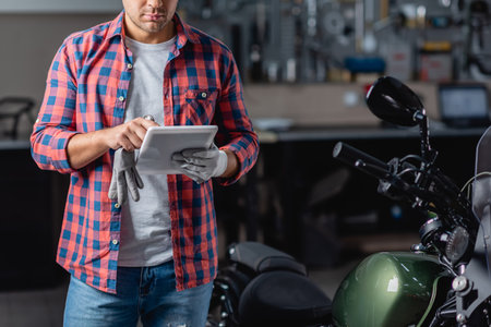 partial view of mechanic in checkered shirt using digital tablet near motorcycle in workshopの写真素材