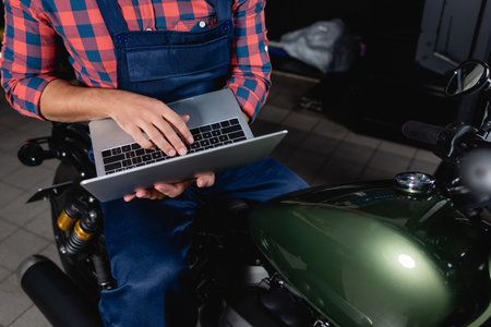 cropped view of technician using laptop while sitting on motorbike in workshopの写真素材