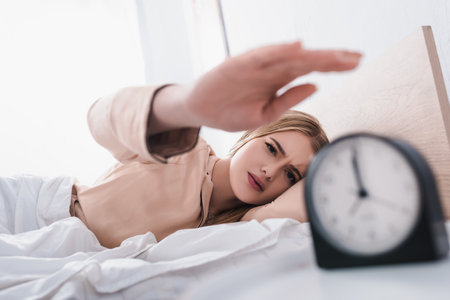 displeased young woman reaching alarm clock on bedside table and blurred foregroundの写真素材