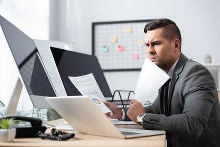 serious trader holding papers near laptop and monitors with blank screen