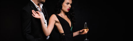 brunette woman in dress holding glass of champagne and pulling away man in suit isolated on black, bannerの写真素材