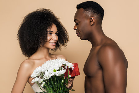 shirtless african american man giving flowers and present to happy woman isolated on beigeの写真素材