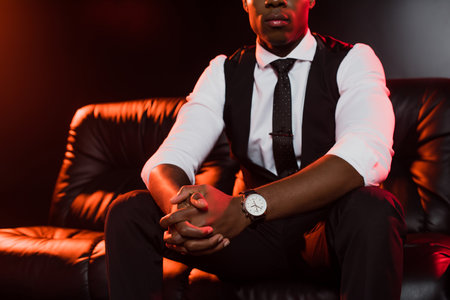 cropped view of african american man in suit sitting on sofa with clenched hands on black backgroundの写真素材