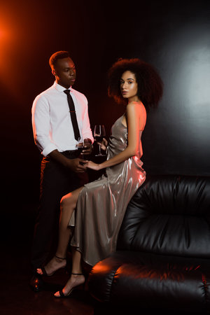 curly african american woman and man holding glasses of red wine near sofa on blackの写真素材