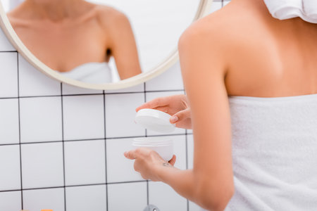 cropped view of woman opening cosmetic cream near blurred reflection in mirrorの写真素材