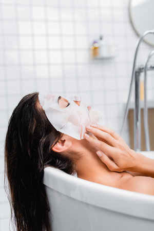 young woman touching face mask while taking bath at homeの写真素材