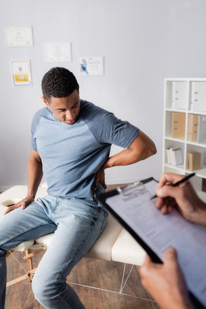 young african american patient suffering from pain in back near doctor writing prescription on...