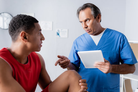 chiropractor holding digital tablet while talking and gesturing near african american patient in...