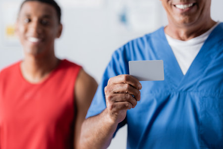 cheerful doctor holding blank card near african american patient in sportswear on blurred backgroundの写真素材
