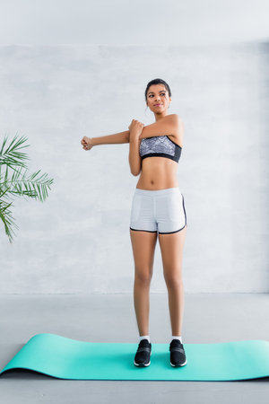 full length view of young african american sportswoman stretching arms on fitness mat at home
