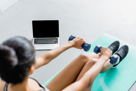 overhead view of african american sportswoman exercising with dumbbells near laptop with blank screen, blurred foregroundの写真素材
