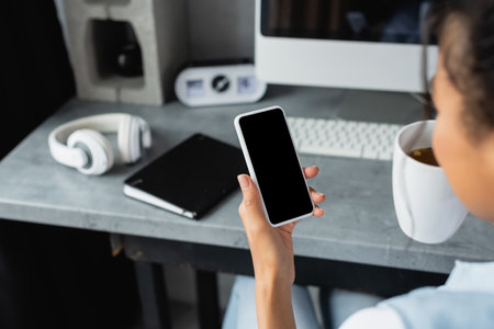 cropped view of african american freelancer holding smartphone with blank screen near notebook and headphones on blurred backgroundの写真素材