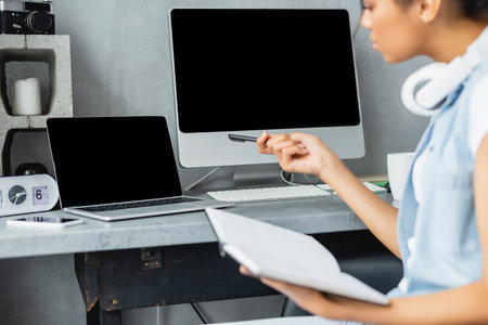 partial view of african american freelancer holding notebook and pointing with pen near laptop and...