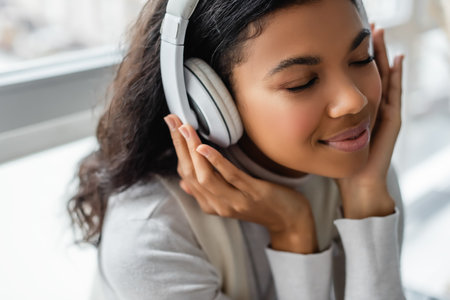 dreamy african american woman listening music in wireless headphones with closed eyes, blurred backgroundの写真素材