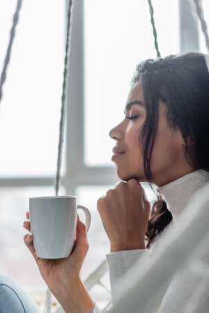 side view of young african american woman holding cup of warm tea with closed eyes, blurred foregroundの写真素材