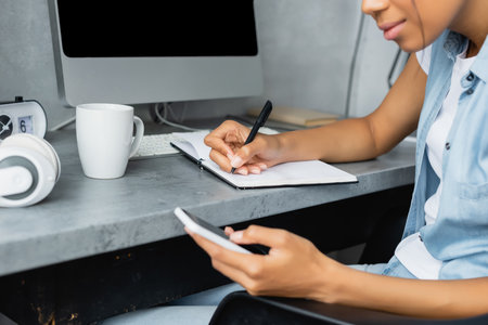 cropped view of african american freelancer using mobile phone and writing in notebook near cup of tea and computer, blurred foregroundの写真素材