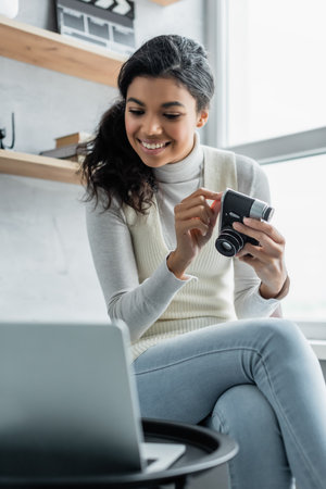 happy african american woman holding vintage photo camera near laptop on blurred foregroundの写真素材