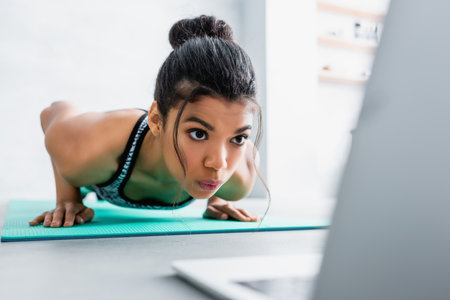 african american sportswoman doing push ups exercise near laptop on blurred foregroundの写真素材