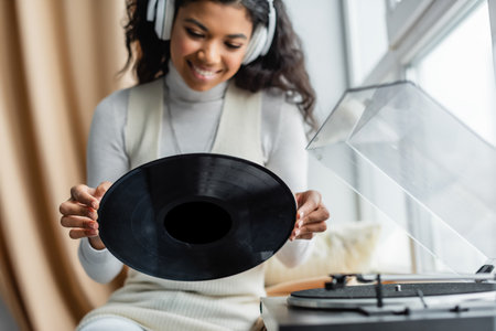 happy african american woman in wireless headphones holding vinyl disc near record player, blurred foregroundの写真素材