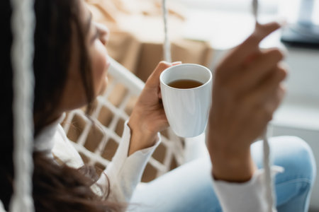 young african american woman resting in hanging chair with cup of tea, blurred foregroundの写真素材