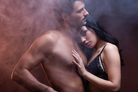 sexy woman leaning on chest of shirtless muscular man while looking at camera on dark background...